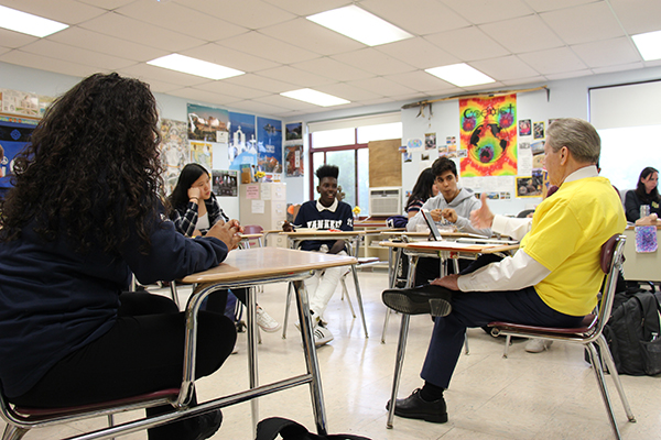 High school students sit in desks arranged in a circle. A man in a white shirt and gold t-shirt over it sits with them as they talk to him.