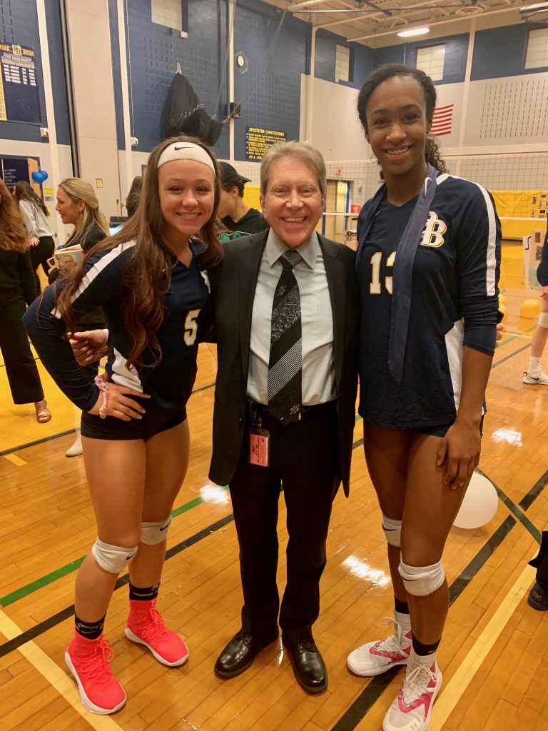 A man wearing a suit and tie is smiling as he stands between two young women in volleyball uniforms.