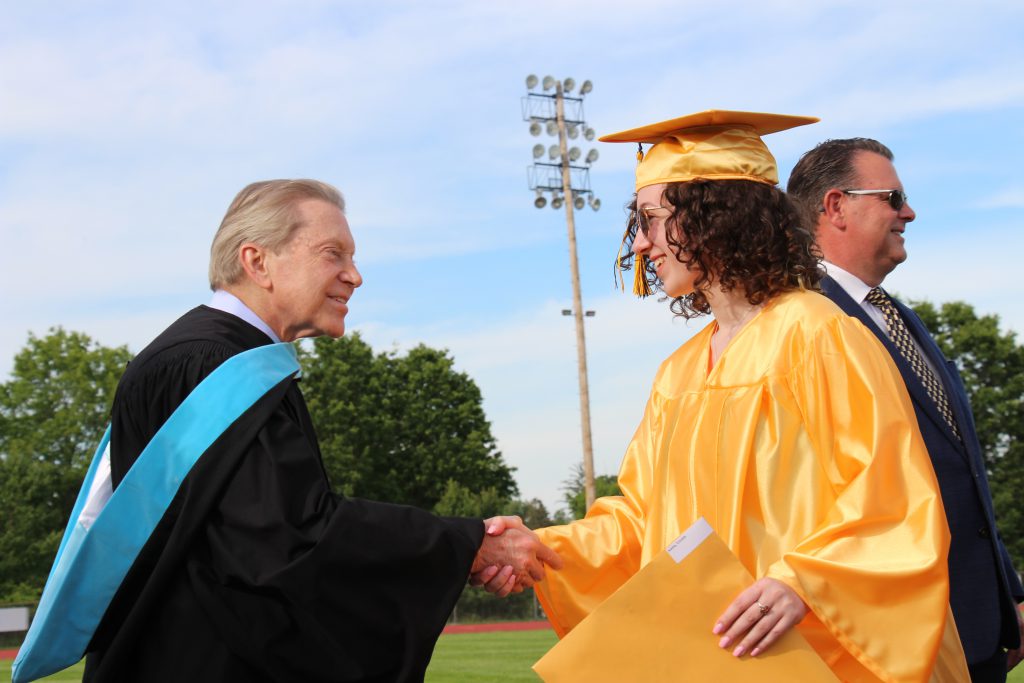 A man wearing a black graduation robe with a blue sash shaking hands with a young women in a gold cap and gown.