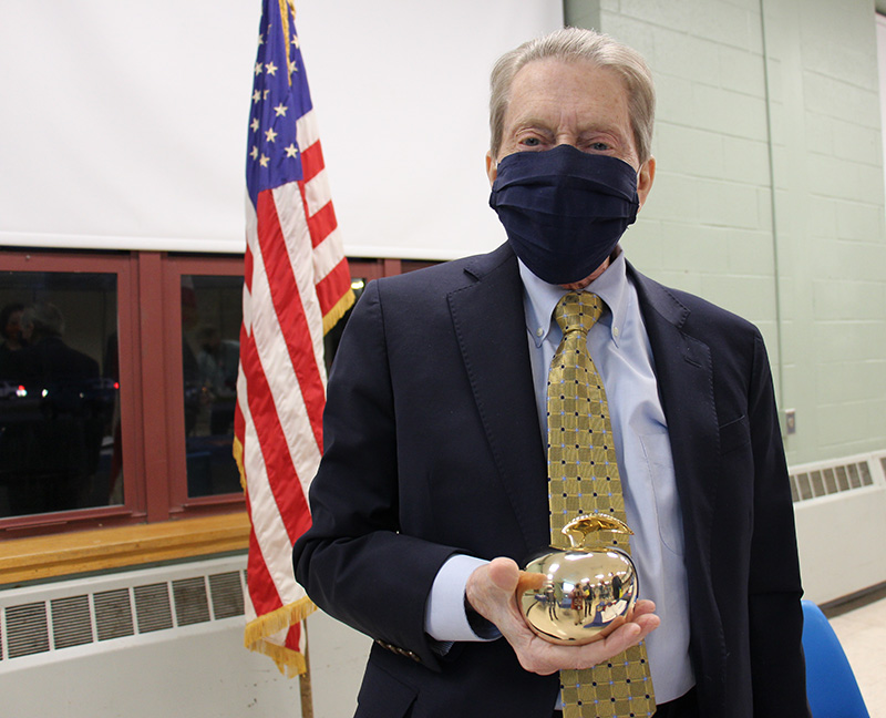 A man in a dark suit, blue shirt and gold and blue striped tie holds a golden apple. 