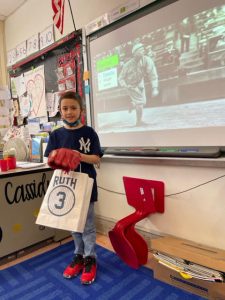A young boy wearing a blue t shirt that has the New york Yankees emblem on it. He is wearing a red  baseball glove and holding a bag that says Ruth with a 3 in a circle. Behind him on a screen is an old time baseball player.