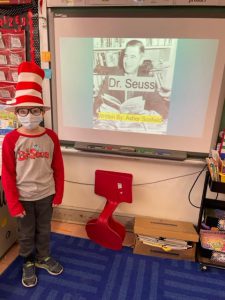 A third grade student stands in front of a screen that has a picture of a man on it. It says Dr. Seuss. The student is wearing a tall red and white striped hat and a long-sleee shirt that says Dr. Seuss