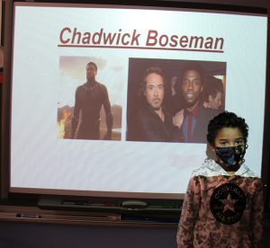 A third grade boy stands in front of a screen that has the name Chadwick Boseman with two pictures of men.