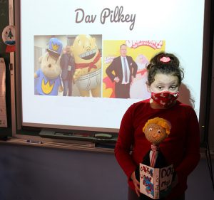 A third grade girl holds a figure of a man in her hands. Behind her is a slide that has photos of a man and characters he created. The name Dav Pilkey is on top.