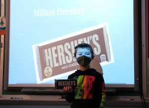 A boy wearing a dark shirt with neon colors on it holds a large Hershey bar. Behind him is a slide projected on a screen with the name Milton Hershey and a picture of a large Hershey bar.