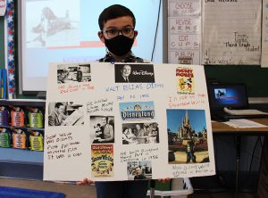 A third grade boy with dark hair holds a poster with all pictures of Walt Disney on it.