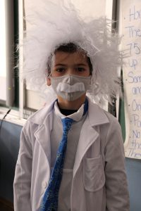 A third grade boy dressed as Albert Einstein, with fluffy white hair, a mask with a white mustache on it, a lab coat and a blue tie.