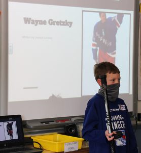 A third grade boy dressed in a NY Rangers jersey, holds a hockey stick. In the background on a screen is a picture of Wayne Gretzky with his name.
