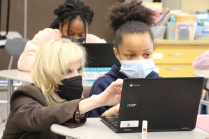 A woman with short blond hair crouches down next to a fifth-grade student and points at her Chromebook. The girl has her dark curly hair pulled back into a ponytail. Both are wearing masks and looking intently at the screen.