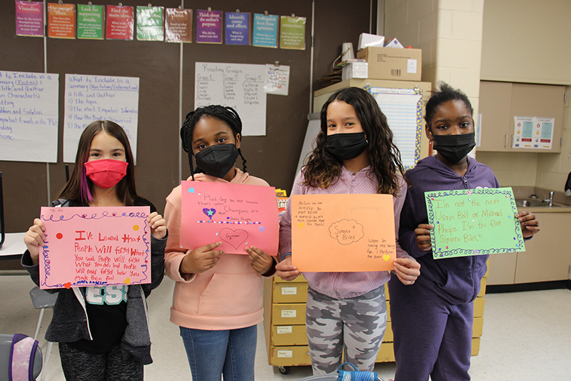 A group of four fifth-grade girl hold their quote pages. They all are wearing masks and have construction paper in front of them. On each sheet, is an inspirational quote written by them from a Black leader in our history.