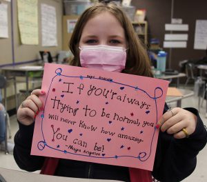 A fifth-grade girl holds up a pink piece of construction paper. On it is written "If you're always trying to be normal, you will never know how awesome you can be. Maya Angelou". There is a blue boarder drawn around it.
