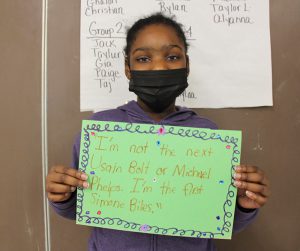 A fifth-grade girl holds a piece of green construction paper up in front of her. There is a fancy blue boarder all around it with multi-color dots here and there. The quote on the page sais "I'm not the next Usain Bolt or Michael Phelps. I'm the first Simone Biles." 
