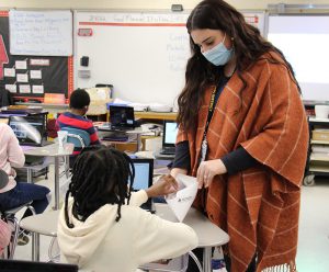 A woman with long brown hair, wearing a blue mask and a long brown poncho holds an envelope out for a student. The student, whose back is to the camera, has multiple braids, is wearing a white hooded jacket and is reaching into the envelope to choose stickers.
