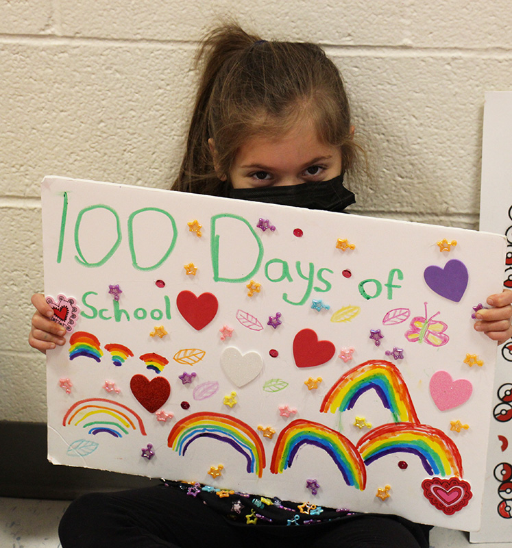 A little girl holds a poster with rainbows on it. It says 100 days of school. There are hearts all around it too.