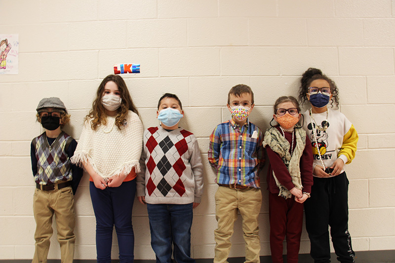 Six first grade students standing in a line. They are all wearing masks and clothes that usually old people wear, like glasses with strings around them, sweater vests.