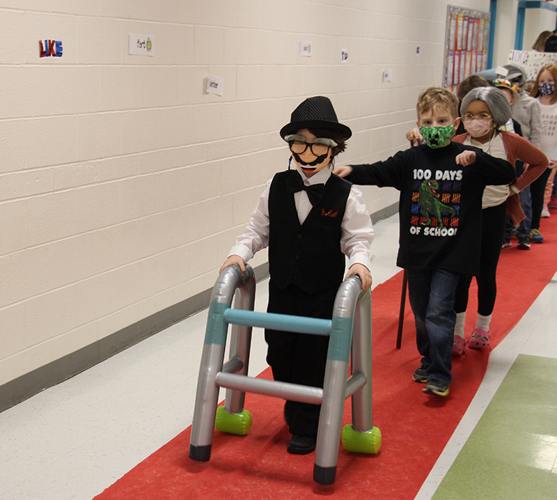 A line of first grade students walking down the hall on a red carpet. First in line is a little boy dressed as an old man, with a suit, hate and a walker.