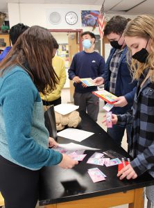 A group of high school students stand around a black table holding small Valentines cards in their hands. Some are writing on them and others are handing them out.