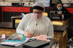 A high school student dressed in a cream colored sweater, wearing glasses, a mask and hair covering, sits at a table and writes out Valentines cards.