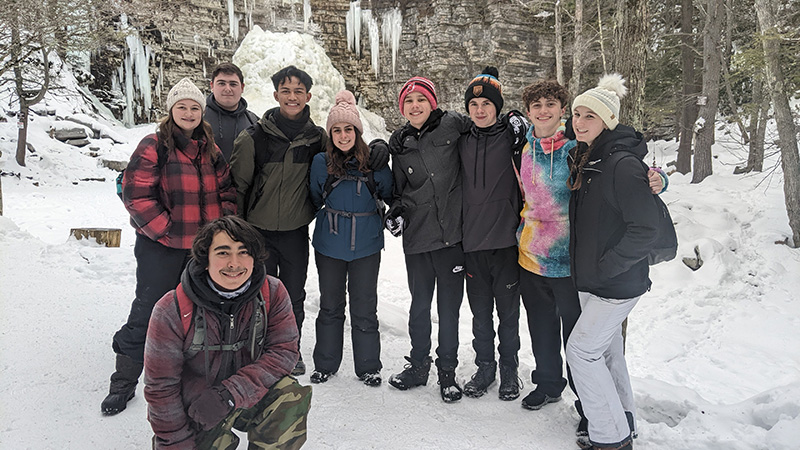 A group of nine high school students stand in front of a stone wall with lots of ice on it.