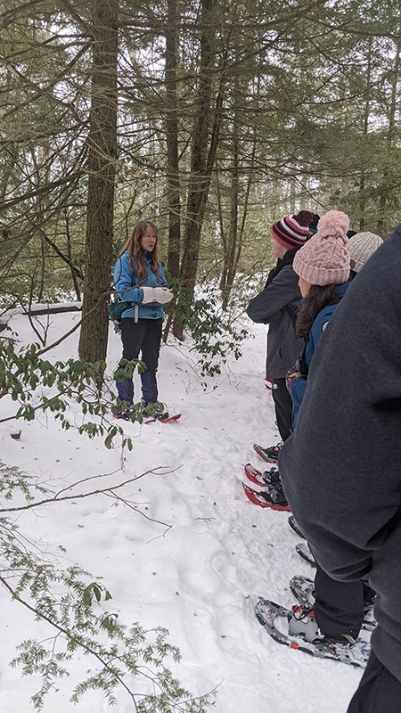 A snowy ground, trees all around. A group of winter coat-clad high school studnets stand and listen to a woman who is wearing mittens and a blue jacket.