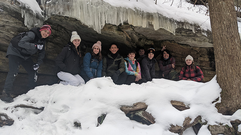 A group of nine high school students stand under a large rock with ice hanging down.