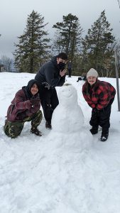 Three high school students stand around a smaller snow man, that has a smile on its face.