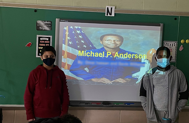 Two middle-school students  on either side of a screen which shows a picture of Michael P. Anderson, a man dressed in blue with an American flag behind him.