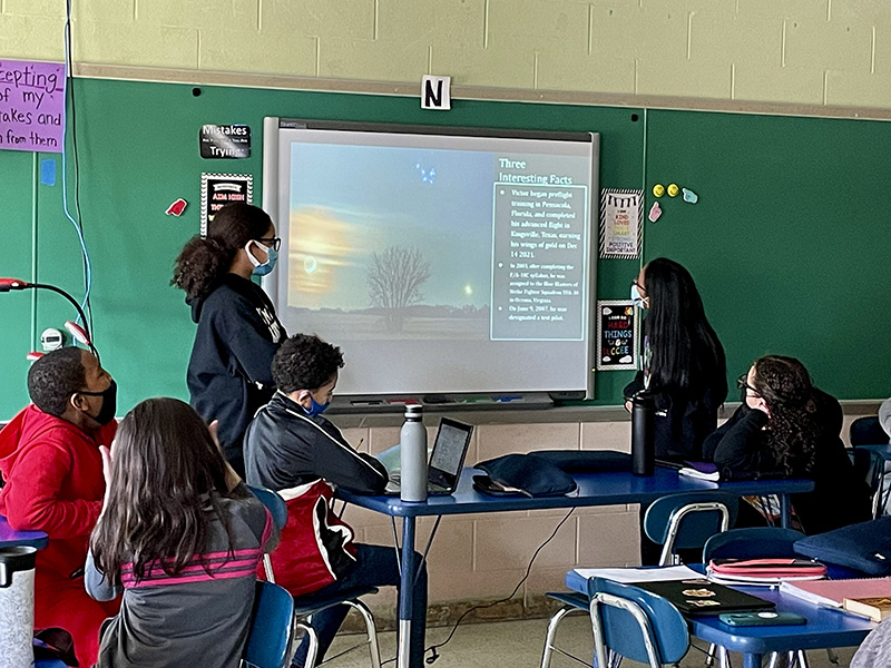 Middle school girls stand on either side of a screen. They are looking at it and describing what's on it to the class.