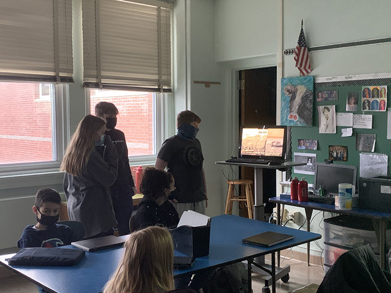 In a classroom setting, a group of three middle school students stand at the front of the class, with a laptop facing the seated students. They are giving a presentation.