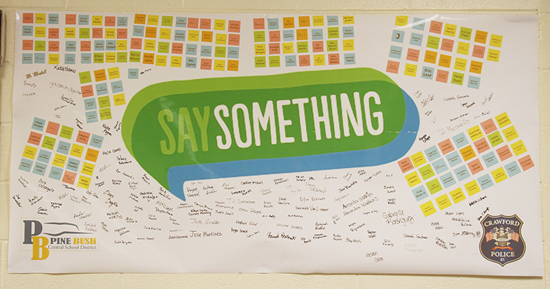 A bulletin board with many different color post-it notes around the top and sides. In the center is a big green word bubble that says Say Something. Many signatures are on it too.