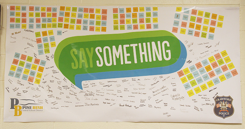 A bulletin board with many different color post-it notes around the top and sides. In the center is a big green word bubble that says Say Something. Many signatures are on it too.
