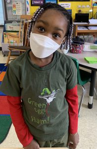 A younger elementary student with long dark hair in briads, wearing a white mask and a gray tshirt with Green Eggs and Ham on it.