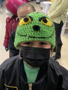 An elementary kid wearing a black jacket, green shirt, black mask and a green knit hat that looks like the grinch.