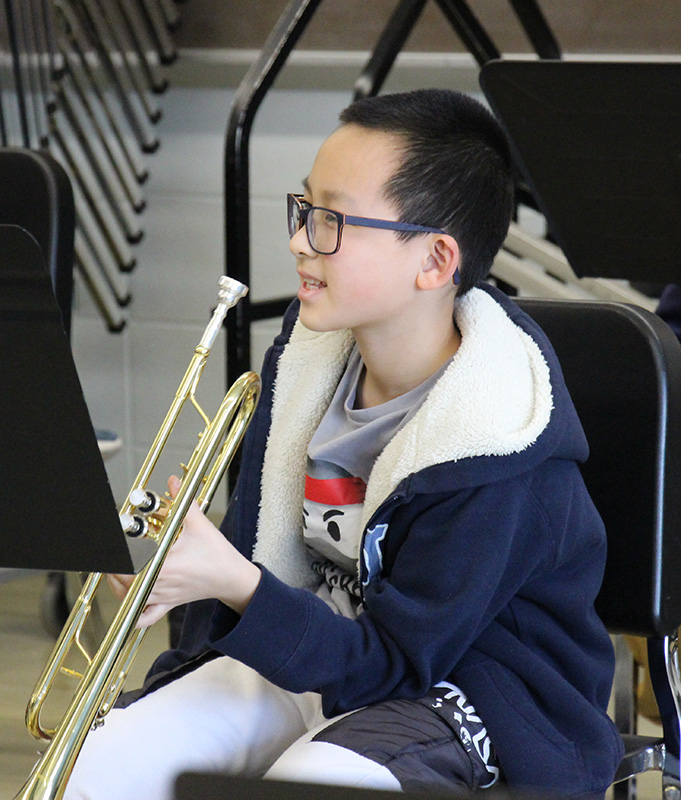 A boy with short dark hair, glasses, wearing a blue hooded sweatshirt is smiling and holding a trumpet close to his mouth.
