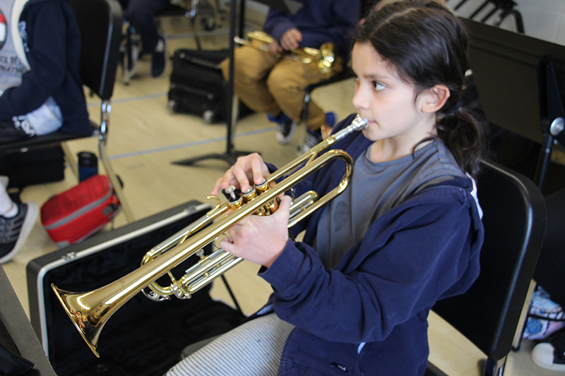 A fifth-grade girl with long dark hair pulled back, wearing a blue sweatshirt, plays a trumpet.
