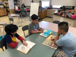 Three elementary students sit at a table reading books.