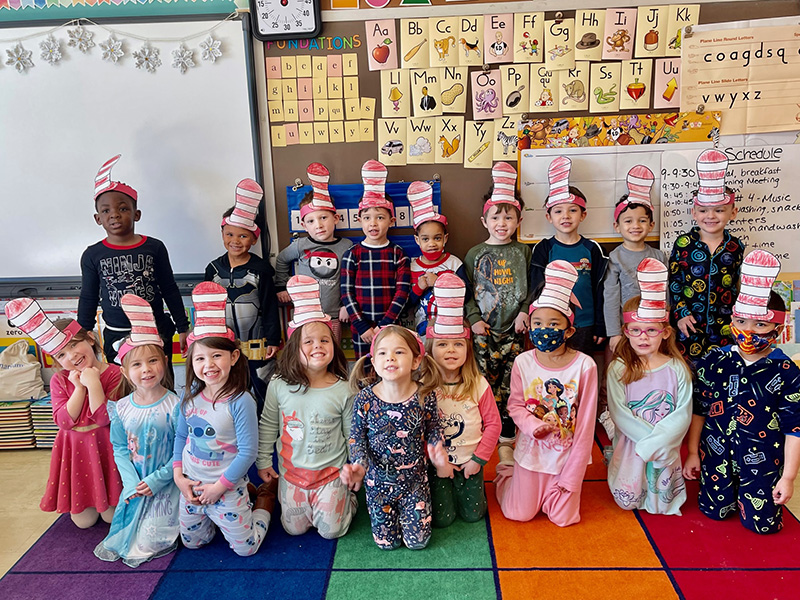 A group of 18 preK students in two rows. First row is kneeling the second is standing. They are all smiling and wearing pajamas! They have tall red and white striped paper hats on their heads. They are all in a classroom.