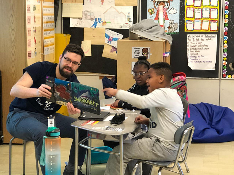 A man with glasses and a book open while three students sitting around the table point at it.beard and mustache holds