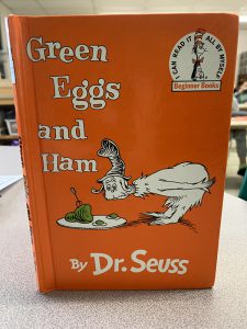 A book with an orange cover. The words Green Eggs and Ham by Dr. Seuss are on it along with a character leaning over a plate that has green eggs and ham on it.