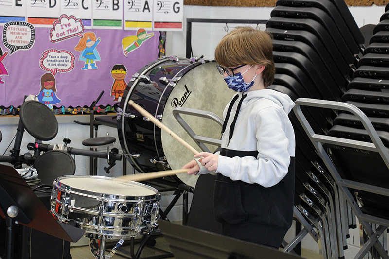 An elementary age boy with shorter blonde hair, wearing glasses, a mask, and a black and white hooded sweatshirt, holds two drumsticks and taps drum.