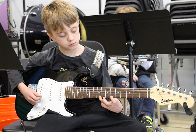 A fifth-grade boy with short blonde hair sits with an electric guitar he is playing.