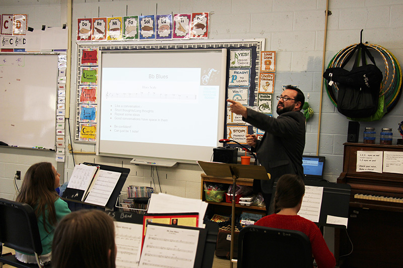 A man points to a screen while students sit in front of him with musical instruments and music stands.
