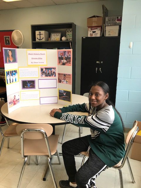 A young woman sits at a table that has a trifold on it with pictures and information. She is smiling.