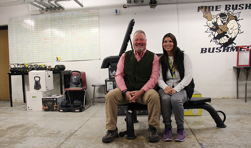 A man wearing a red shirt and blue vest and a woman in a white shirt and gray  sweat pants sit on a weight bench smiling. Behind them is brand new weight equipment.