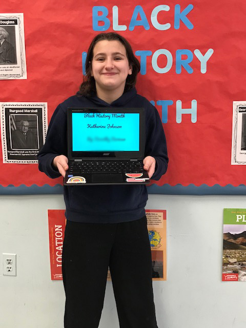 A student smiles in front of a red bulletin board. He is holding a chromebook with his project pulled up. She is smiling.
