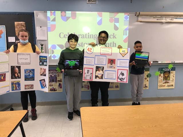 Four students standing in front of a screen. Two of the students are holding trifold boards with photos and information on them. The other two are holding chromebooks with their presentations showing.