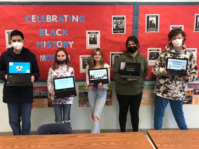 A large red bulletin board in the background that says Celebrating Black History Month in white and blue. In front of the board are five students holding chromebooks with projects on them.