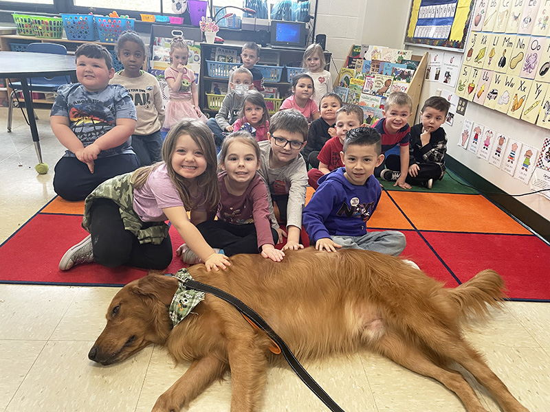 A group of 16 pre-school children sitting on a colorful run. In front of them is a Golden Retriever lying down. Four kids are petting him.