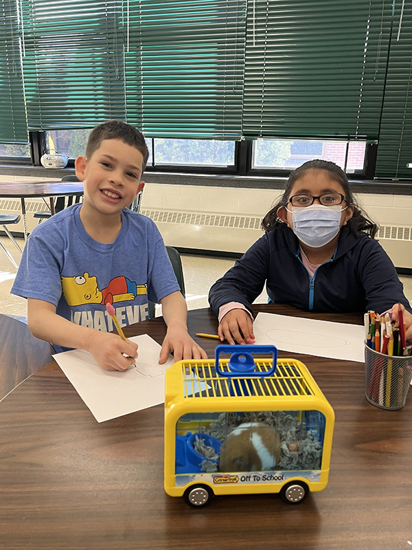 Two elementary students sit at a table with a small cage with a hamster in it. There is a boy on the left smiling and a girl on the right, wearing a mask.