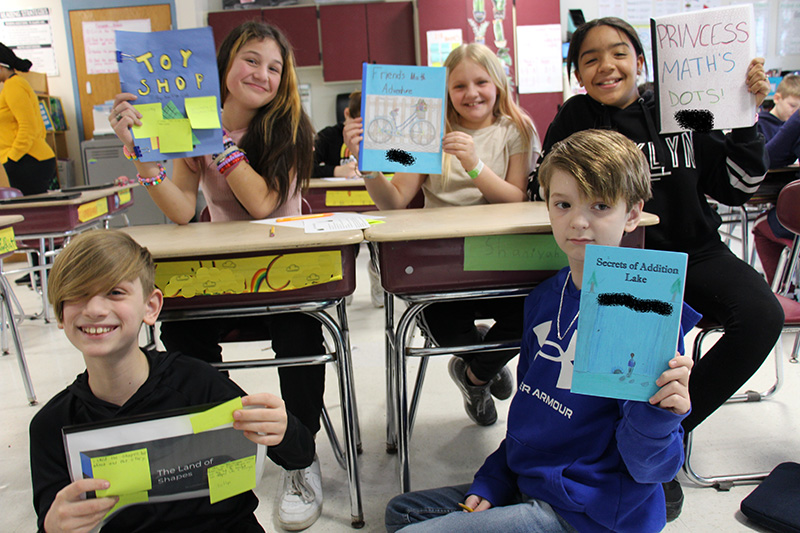 A group of five fifth grade students - two sitting on the floor and three in desks - all holding homemade books about math. They are all smiling.